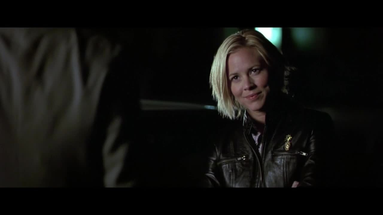 A woman with short blonde hair smiles, looking at someone just out of frame. She's wearing a leather jacket with a small pin on the lapel, the lighting dim around them.
