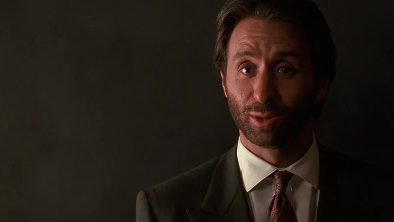 The man in the suit is speaking, his eyes focused and his expression thoughtful. The lighting is dim, casting a shadow behind him.
