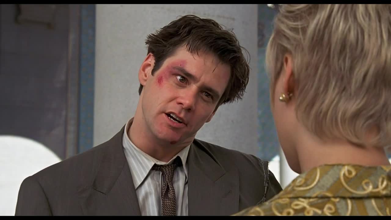 He's speaking, a bruise blooming on his face, and his eyes are locked on the woman in front of him. Her back is to us, and the collar of her patterned jacket is visible.
