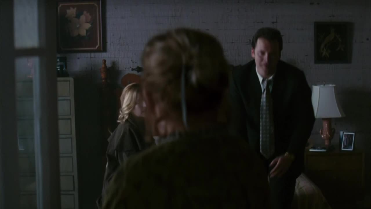A man in a suit stands near a bed, while a woman with blonde hair is partially visible behind someone in the foreground. The lighting is dim, and the scene feels tense.

