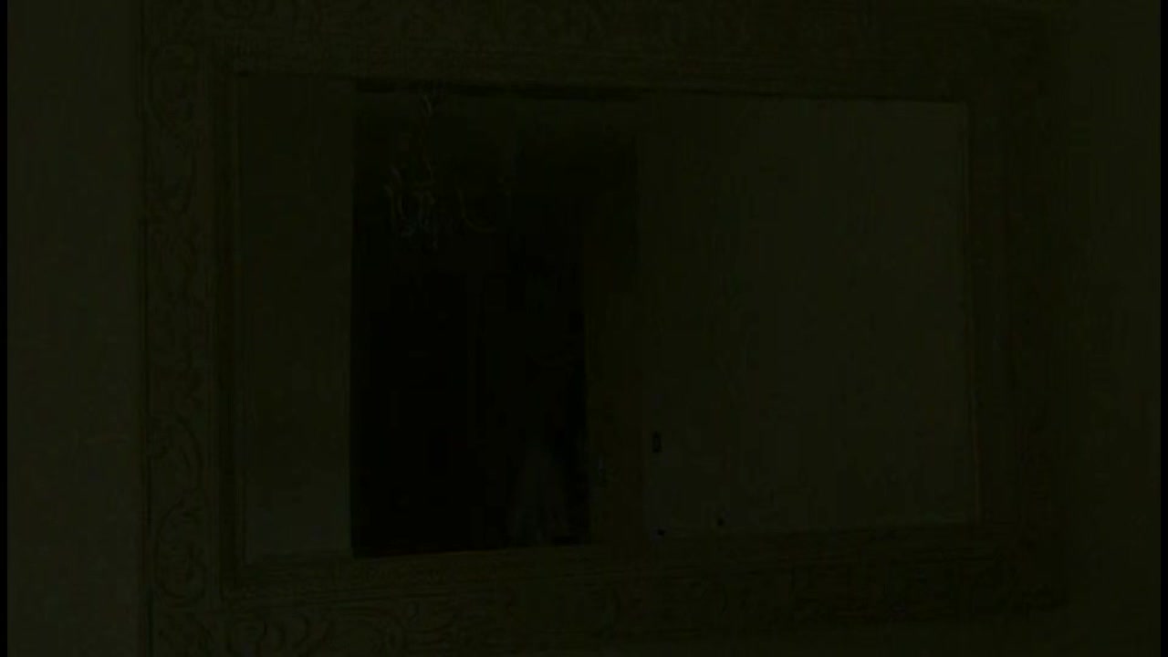A large, ornate mirror hangs on the wall, reflecting a dark room. The frame's intricate carvings are barely visible in the dim light.

