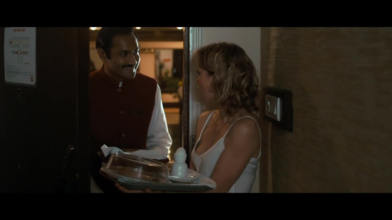 A hotel employee, in a red vest, smiles as he offers a tray with a covered cake and an egg cup to a woman in a white top. She seems to be standing just inside a doorway.
