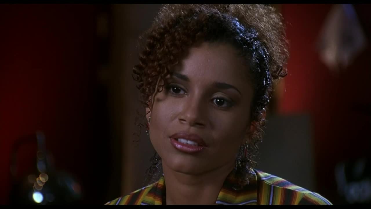 Her lips are slightly parted as she speaks, her gaze directed just off-screen. The light catches the curls in her hair, and the colorful pattern of her shirt is visible.
