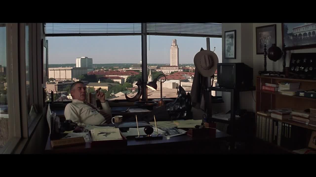 A man in a white shirt and tie leans back in his chair, a lit cigar in his hand. The view outside his office window includes a tall tower and a sprawling cityscape, likely somewhere in the United States.

