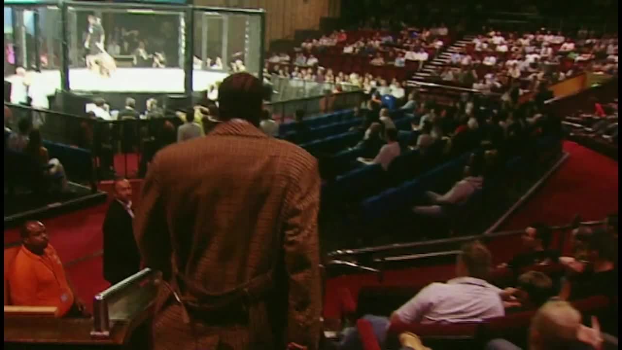 A man in a brown patterned coat walks down the aisle, away from the cage.  Across the arena, the crowd is watching a fight on a large screen.
