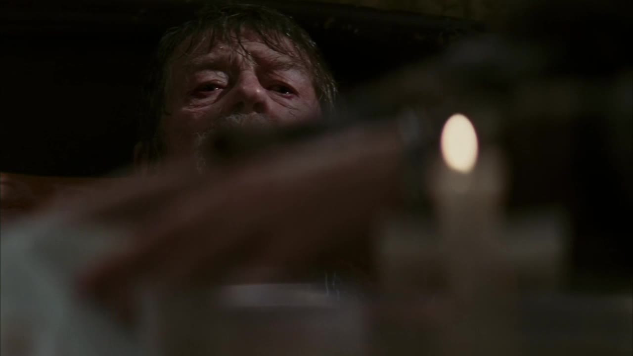 An older man's face is illuminated by a single candle, his eyes fixed on something out of view. A blurry hand reaches into the frame, partially obscuring the man's gaze.
