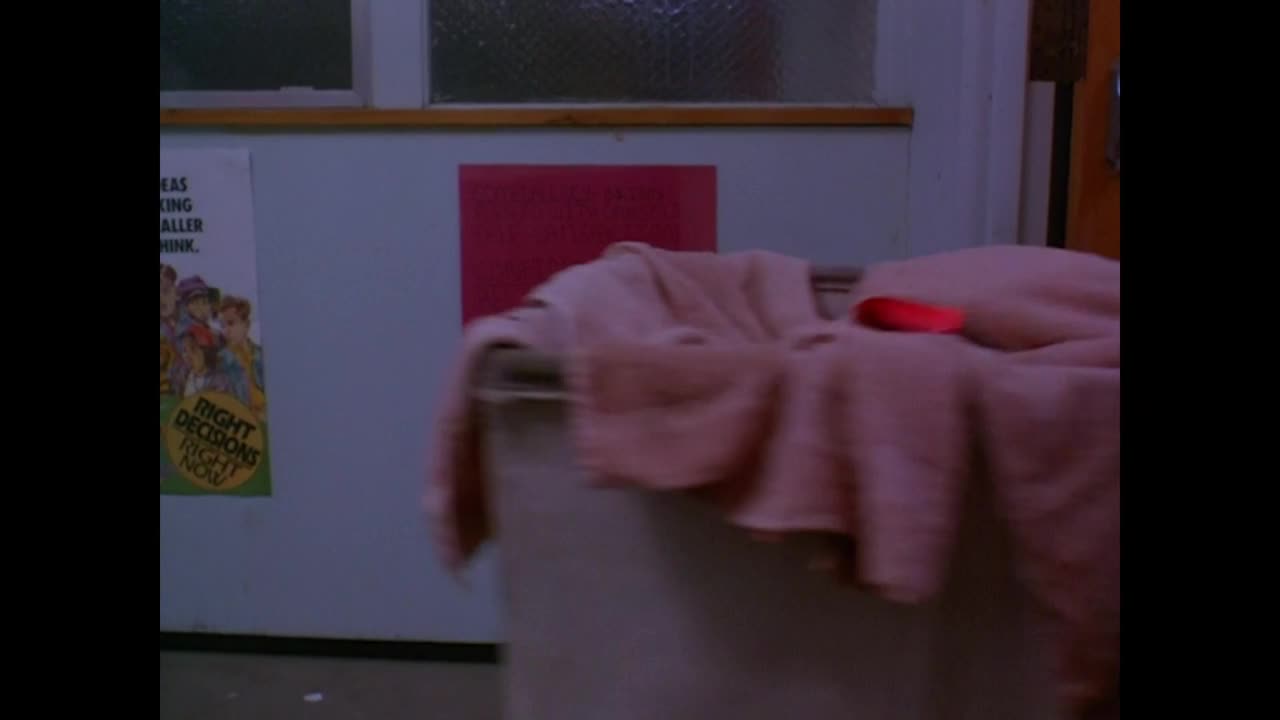A laundry cart, draped with a pink towel, sits in a brightly lit room. A small, red object rests on top of the towel.
