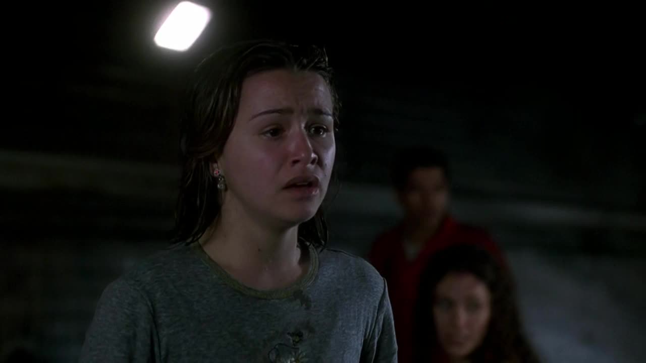 A young woman stares ahead, her face streaked with tears, under a stark, overhead light. Two figures stand behind her, looking on with concern.
