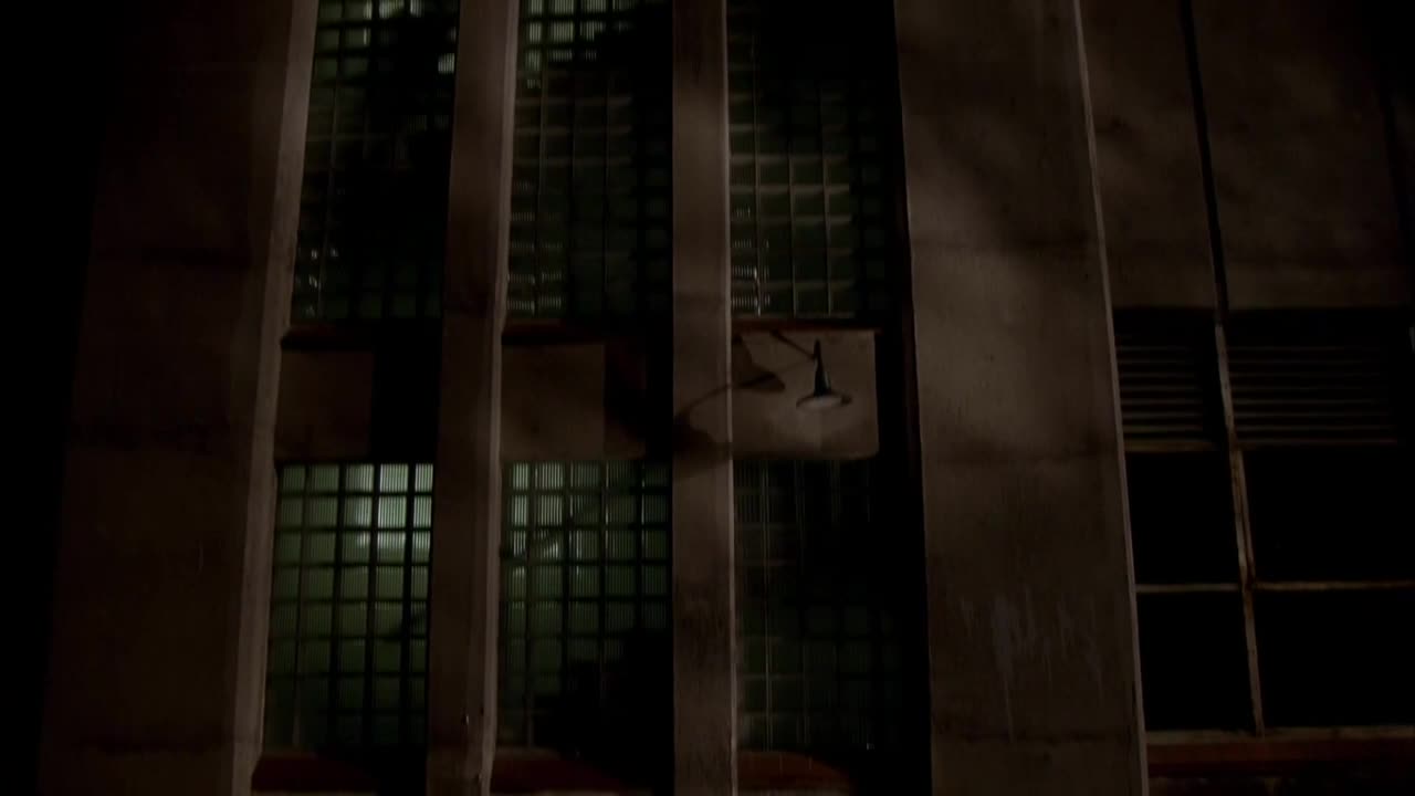 A tall, concrete building looms, its windows filled with a grid of glass blocks. A single, industrial-style light fixture hangs from the wall, casting a dim glow.
