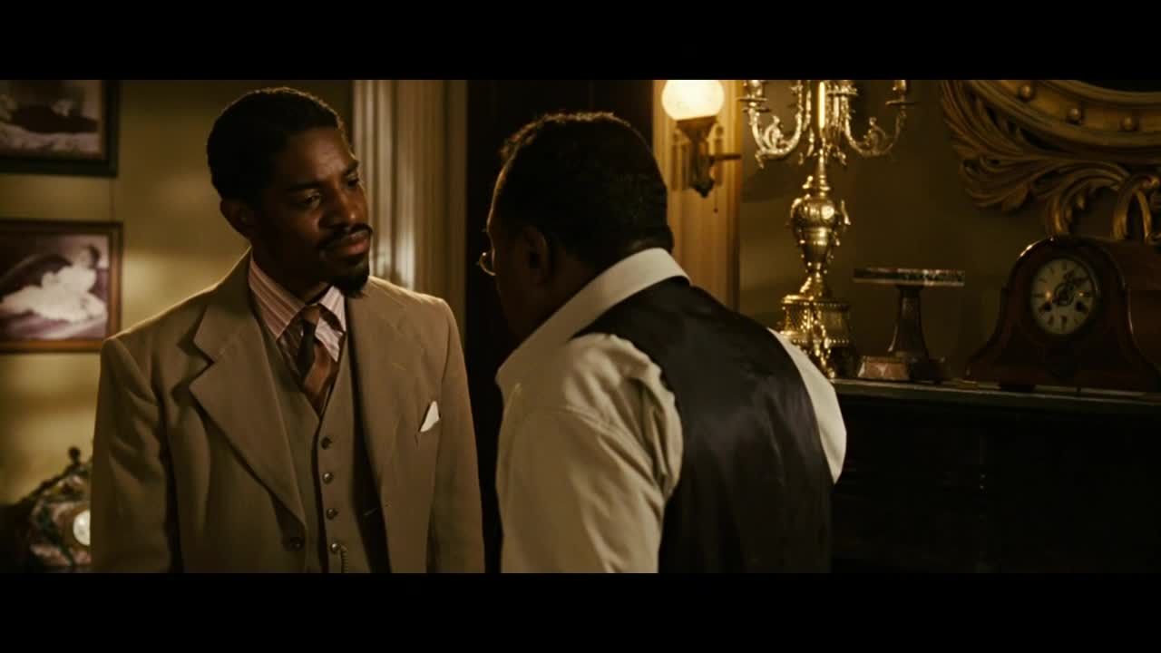 A man in a tan suit listens intently as another, wearing a black vest, speaks. The scene, lit by a warm lamp, takes place in a room with ornate decor and a grandfather clock.
