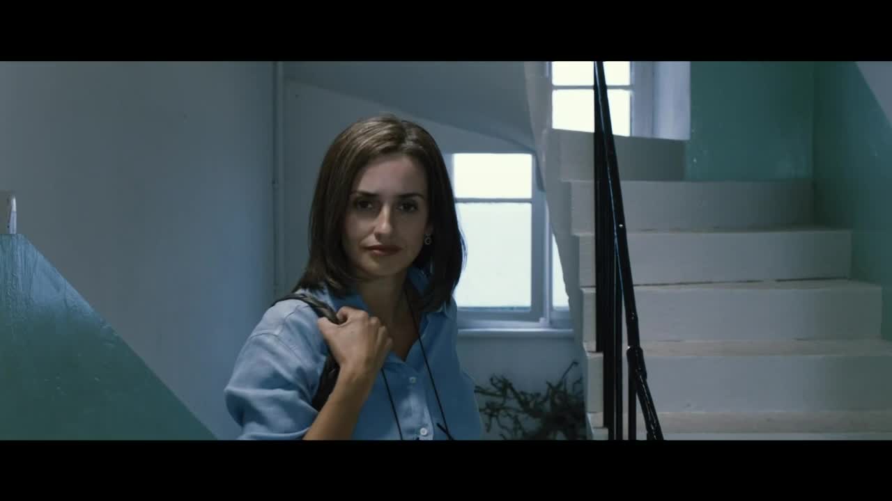 A woman in a blue shirt, with a backpack slung over her shoulder, looks directly at the camera. She stands in a stairwell, with a window visible behind her.
