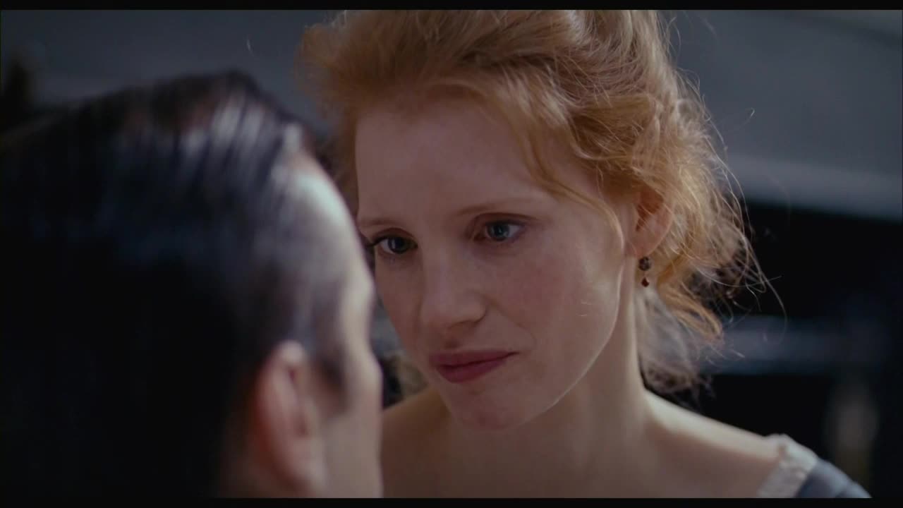Her gaze is locked on the man in front of her, her expression a mixture of curiosity and something deeper. The light catches the red in her hair and the delicate lace at her neckline.
