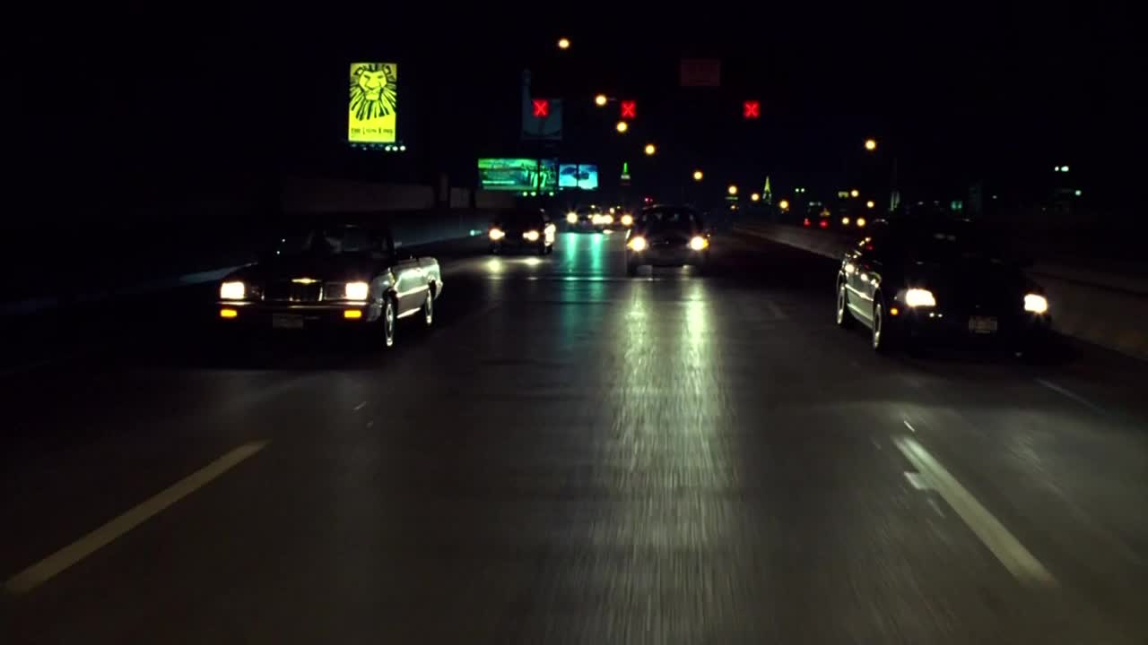 Headlights cut through the darkness as cars travel down the highway. A convertible with its top down leads the way, its front end illuminated by the streetlights.
