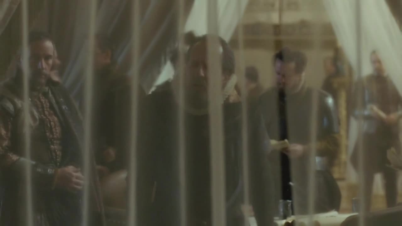 Men in period clothing mill about, some holding papers, while others stand and watch. The scene appears to be indoors, perhaps a meeting or planning session, from a Starz Encore Classic production.

