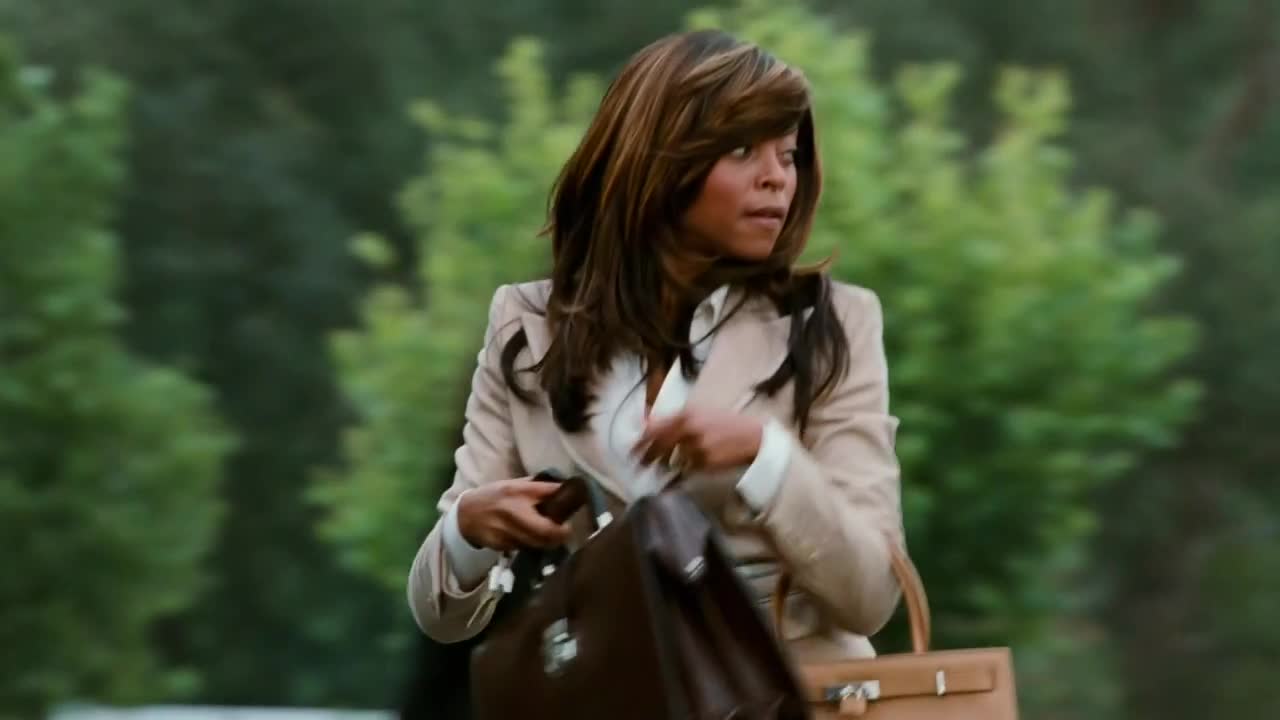 She's hurrying along, clutching two bags, her gaze fixed over her shoulder. The wind is catching at her hair as she moves through the blurred green background.
