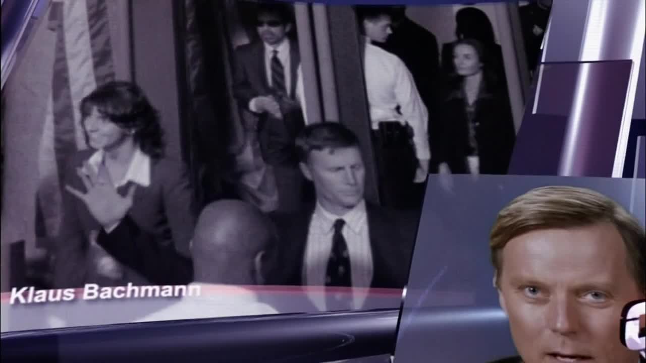 A woman in a blazer gestures with her hand as a group of people in suits and ties move past. In the corner, a man with blond hair looks directly at the camera.
