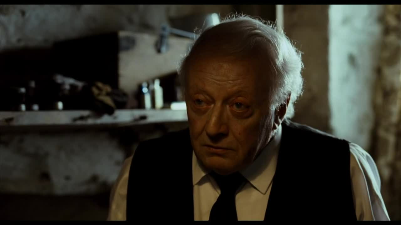 The man stares downward, his face etched with concern. His white hair contrasts with the dark vest and tie he wears, as he sits in a dimly lit room.
