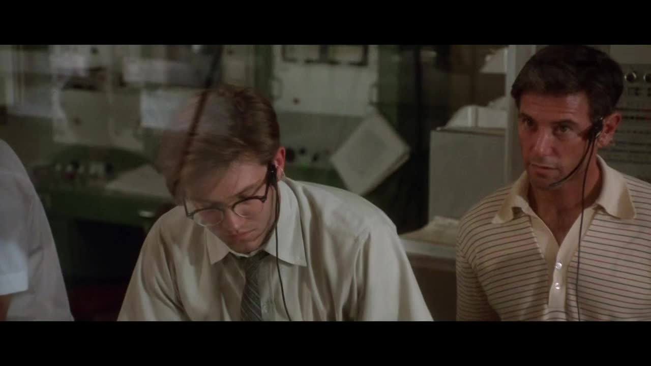 A man in glasses concentrates on something on the desk, while another, in a striped shirt, looks on with a serious expression. Both wear headsets, and the room is filled with equipment.
