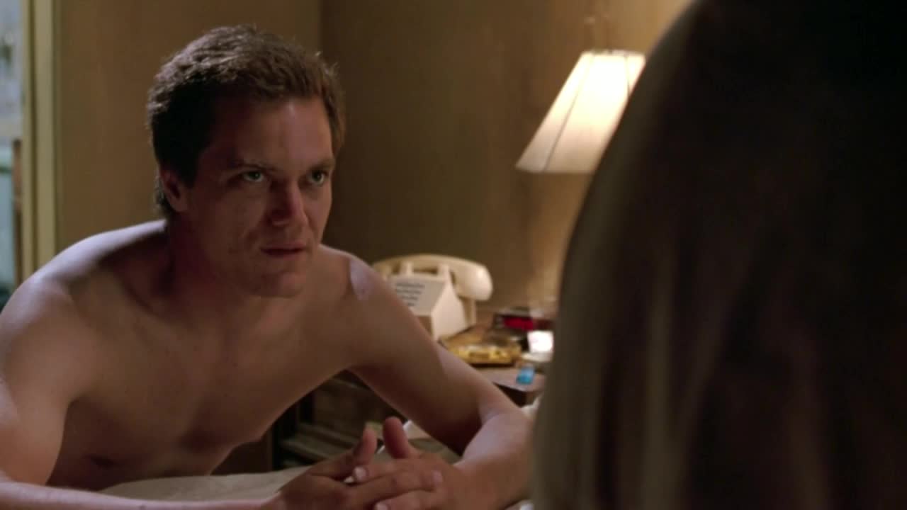 A shirtless man with tousled hair stares intently at someone just out of view. The warm light from a bedside lamp illuminates the room as he leans forward, hands clasped.
