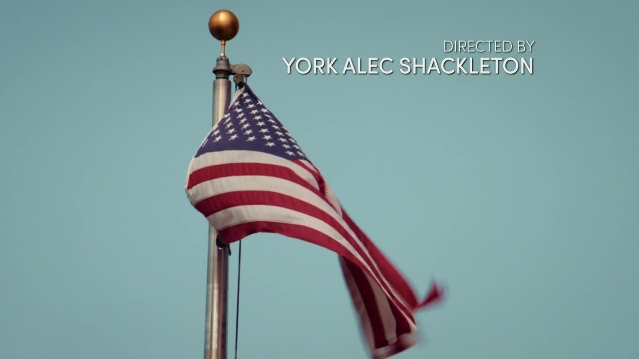 The American flag whips in the wind, its red and white stripes rippling against a pale blue sky. The gold finial atop the pole glints in the light.
