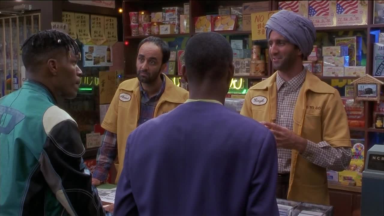 Two men in yellow work shirts stand behind a counter, talking to two customers. The man with the turban gestures with his hands as he speaks.
