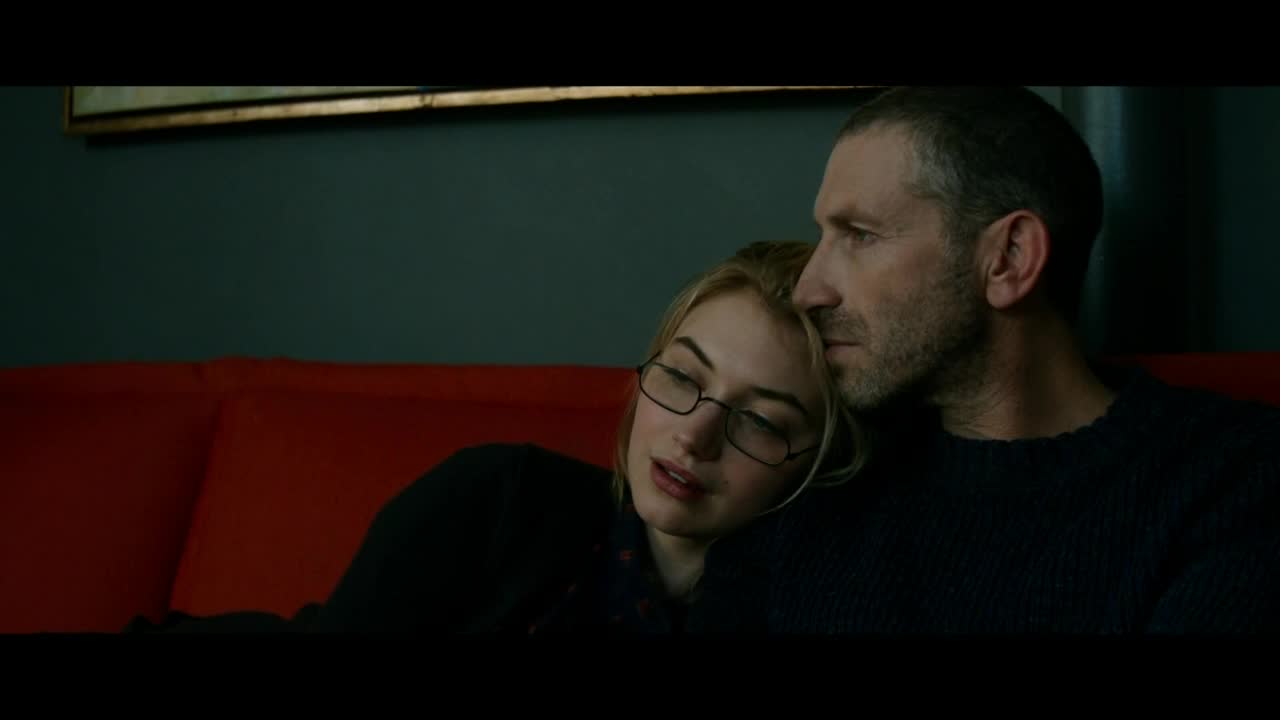 On a red couch, a woman rests her head against a man's shoulder. He looks off to the side, while she wears glasses and appears to be drifting off.
