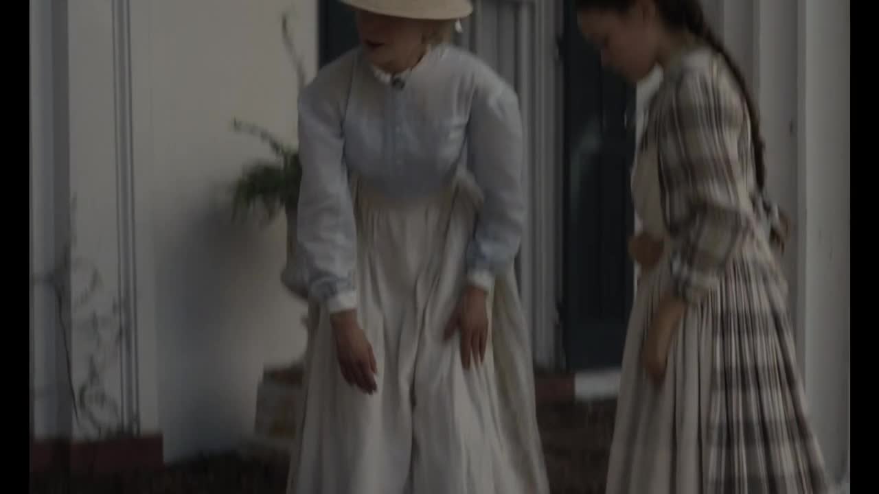 A woman in a light blue dress and a straw hat stands with her hands at her waist, looking down. Beside her, another woman in a plaid dress bows her head.
