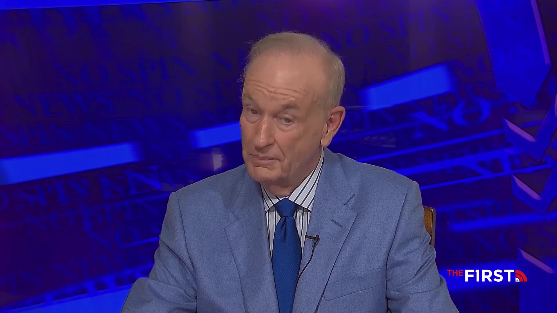 A man in a light blue suit sits at a table, his gaze directed off-camera. The backdrop is a dark blue, and the logo for The First TV is visible in the corner.
