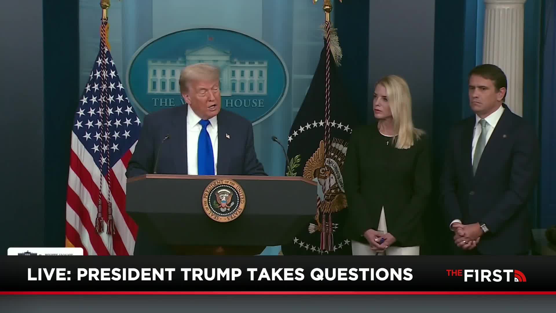 The President, wearing a blue tie, speaks from behind a podium adorned with the Presidential Seal. Two people stand to his right, watching, as a broadcast from The First TV rolls live.
