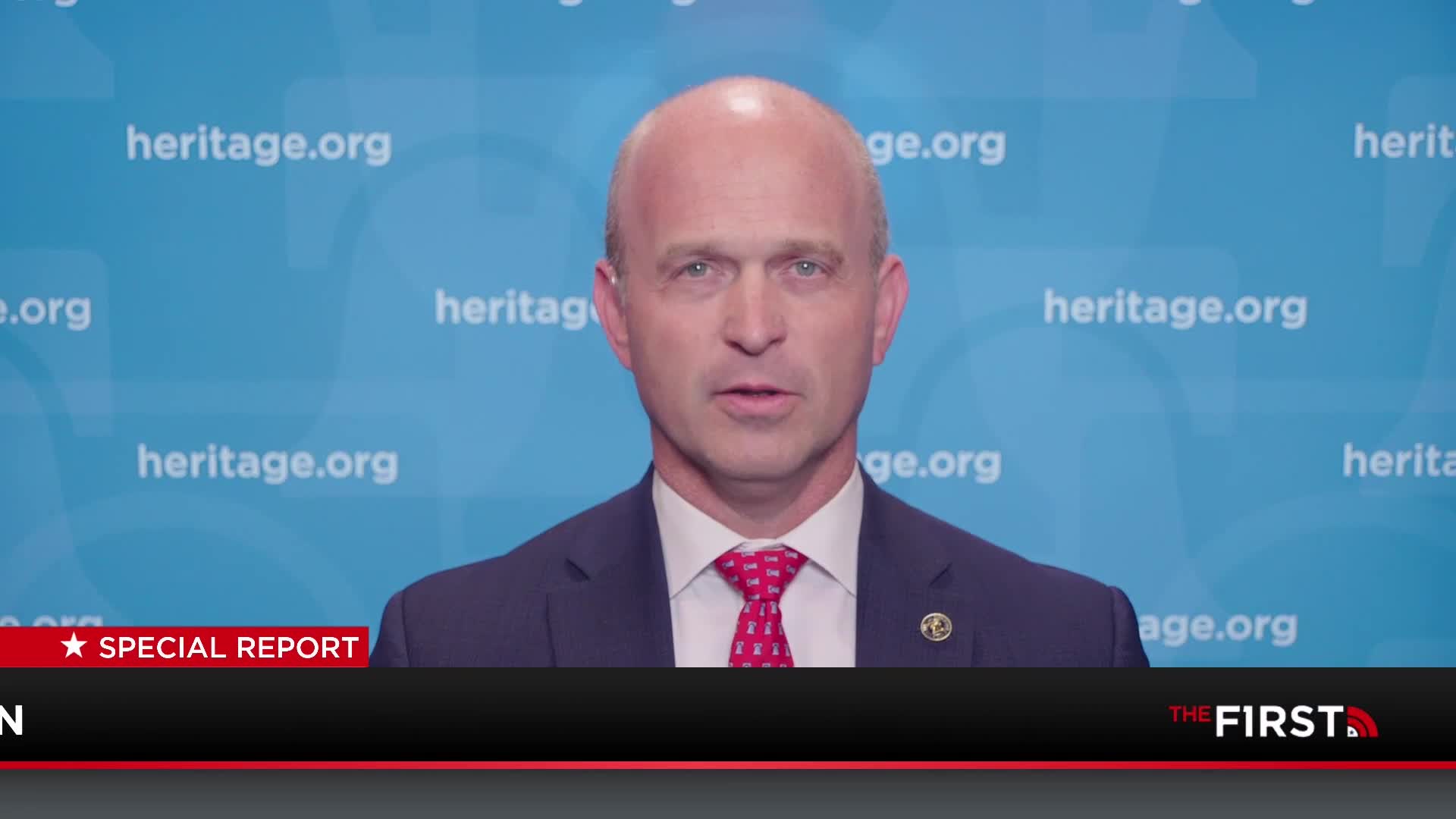 A man in a suit and tie is speaking, his face centered against a light blue backdrop with "heritage.org" repeated. The bottom of the screen displays "THE FIRST" in a red and black banner, indicating a broadcast from a United States TV channel.
