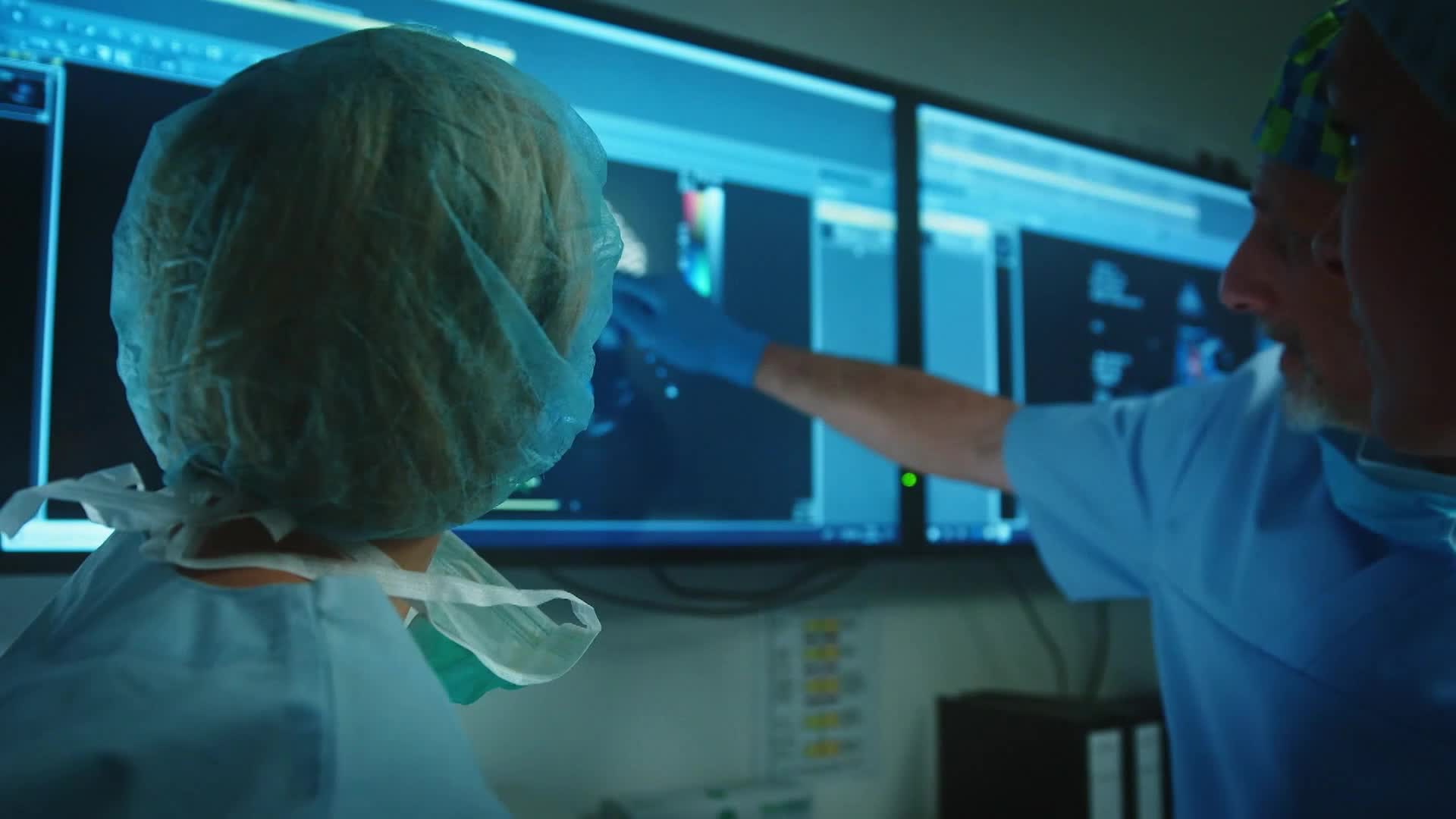 A doctor in blue gloves gestures at a screen displaying medical images, while another person, also in scrubs, looks on. The monitors glow with a cool blue light, reflecting off the surgical caps.
