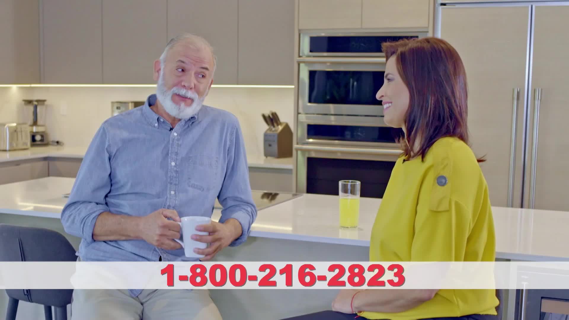 A man with a white beard holds a mug, smiling at a woman in a yellow shirt. The woman is sitting at a kitchen island, facing him, with a glass of juice beside her.
