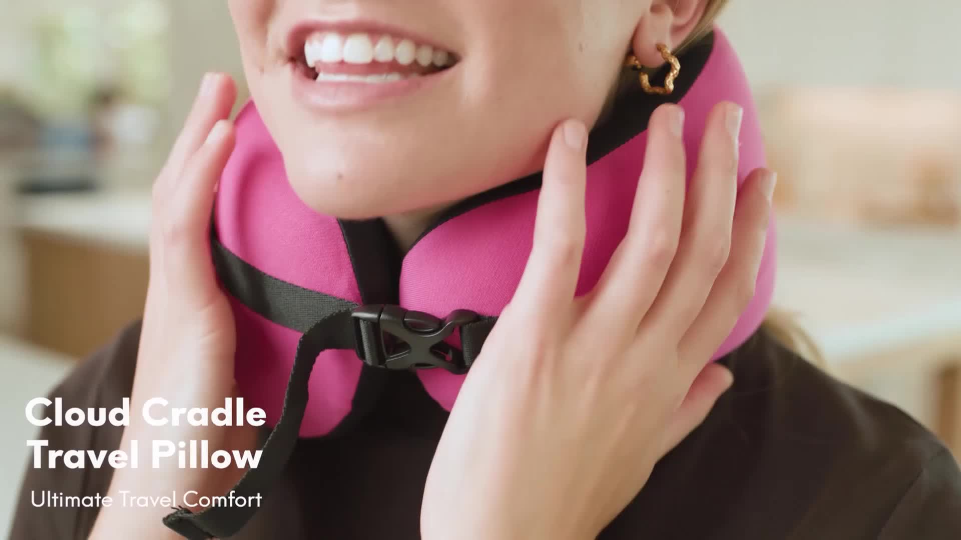 A woman is holding a bright pink travel pillow around her neck, smiling as she adjusts it. The pillow is secured with a black strap and buckle.
