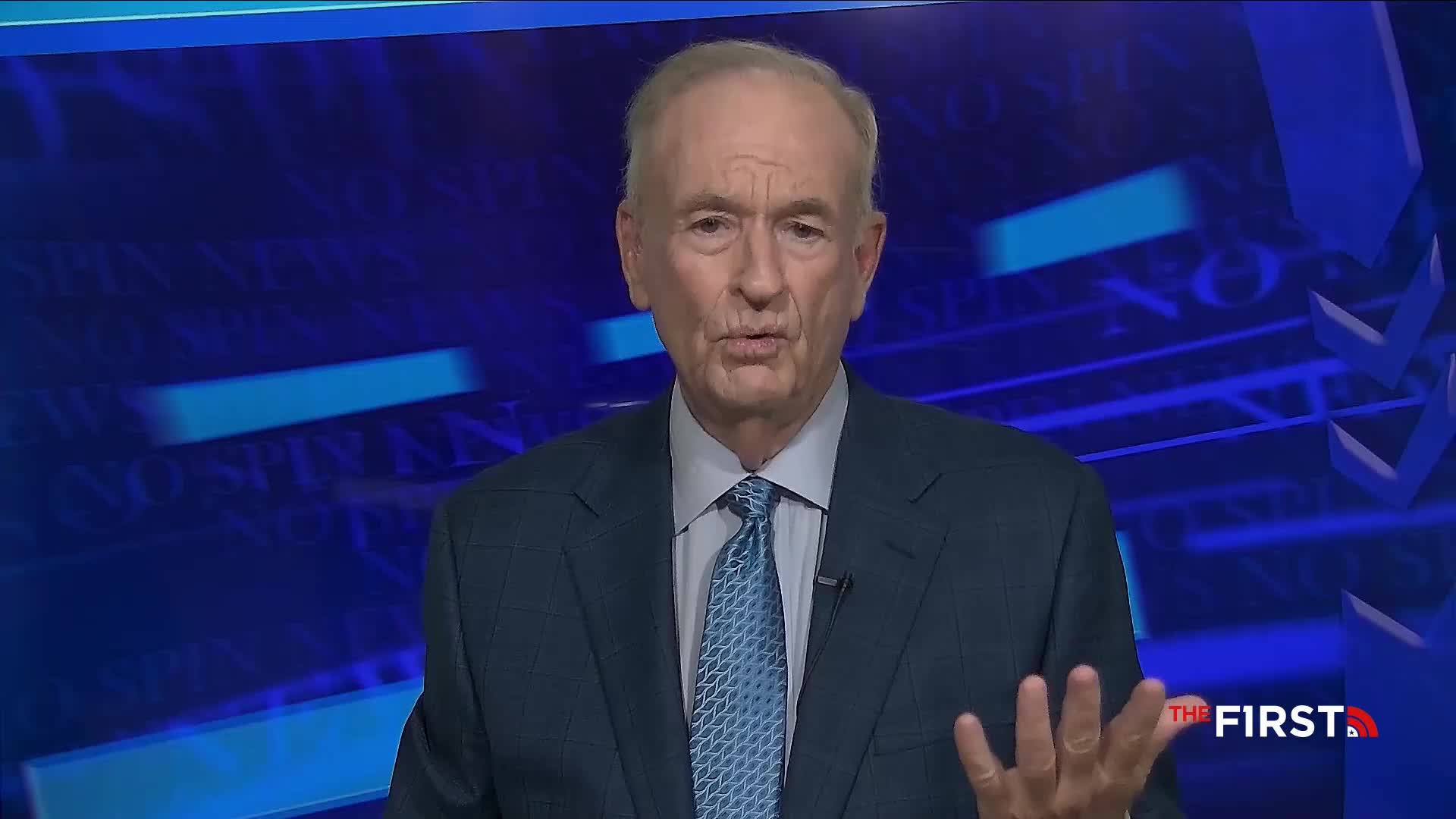 A man in a suit and tie gestures with his hands, speaking directly to the camera. The backdrop is a blue screen with text, and the logo for "The First" is visible in the corner.
