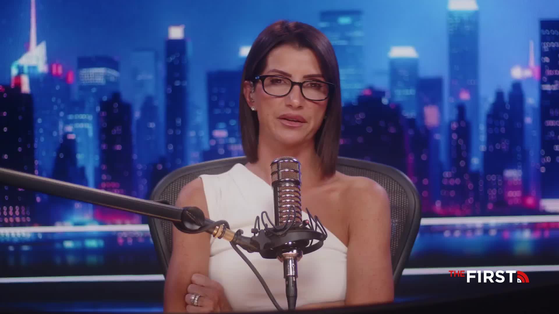A woman wearing glasses and a white top speaks into a microphone, with a backdrop of a city skyline. The logo for "The First" is visible in the lower right corner.
