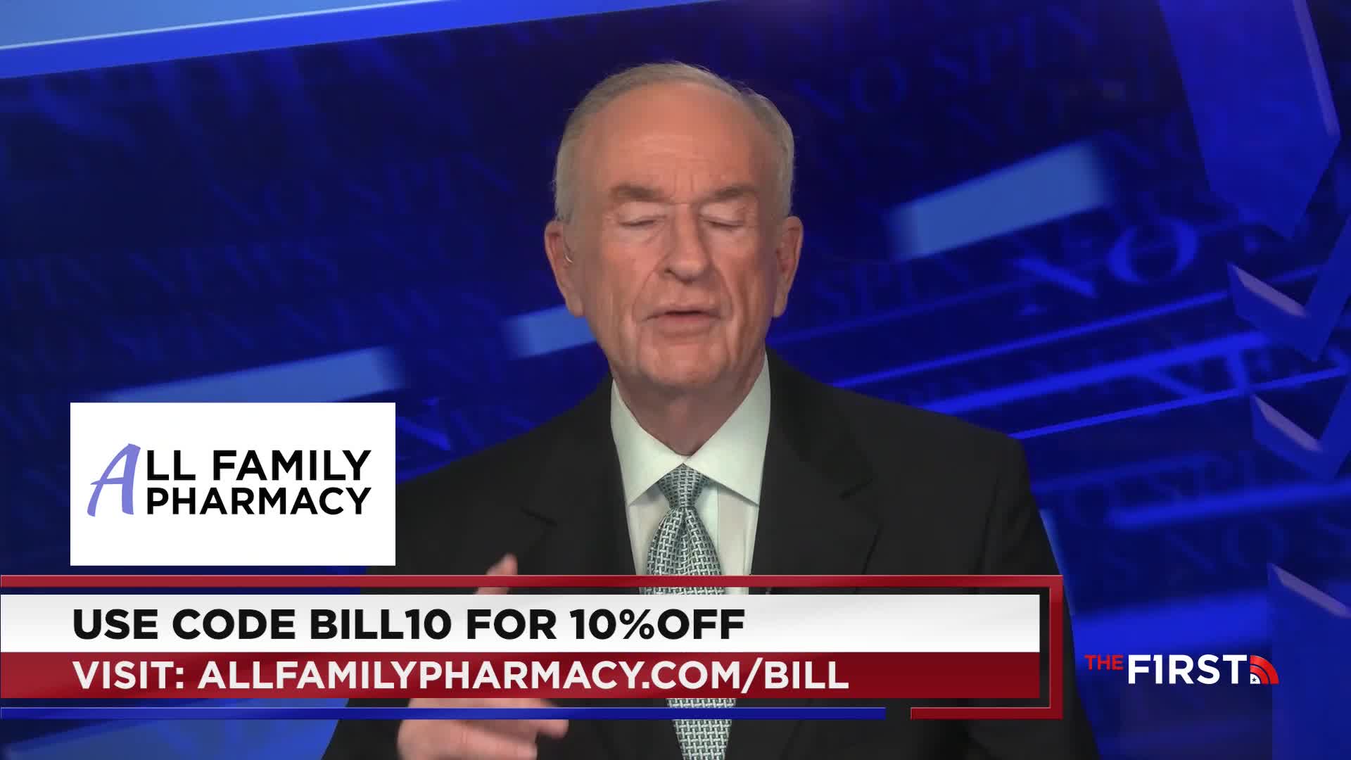 A man in a suit is speaking, his hands gesturing as he talks. Below him, a banner promotes a pharmacy with a discount code, all against a blue backdrop.
