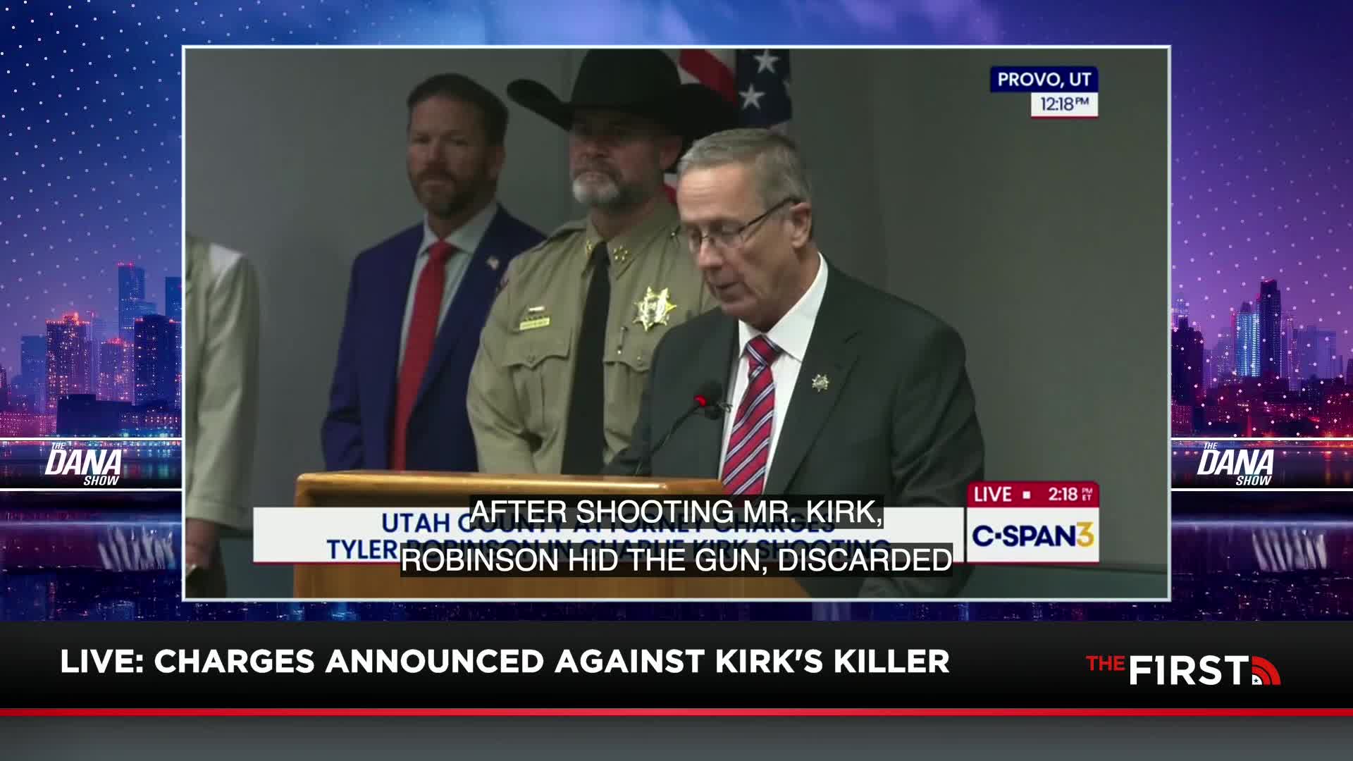 A man in a suit and tie reads from a podium, his face illuminated by the lights. Behind him, a sheriff in uniform and another man in a suit stand stoically, as the charges are announced on The First TV.
