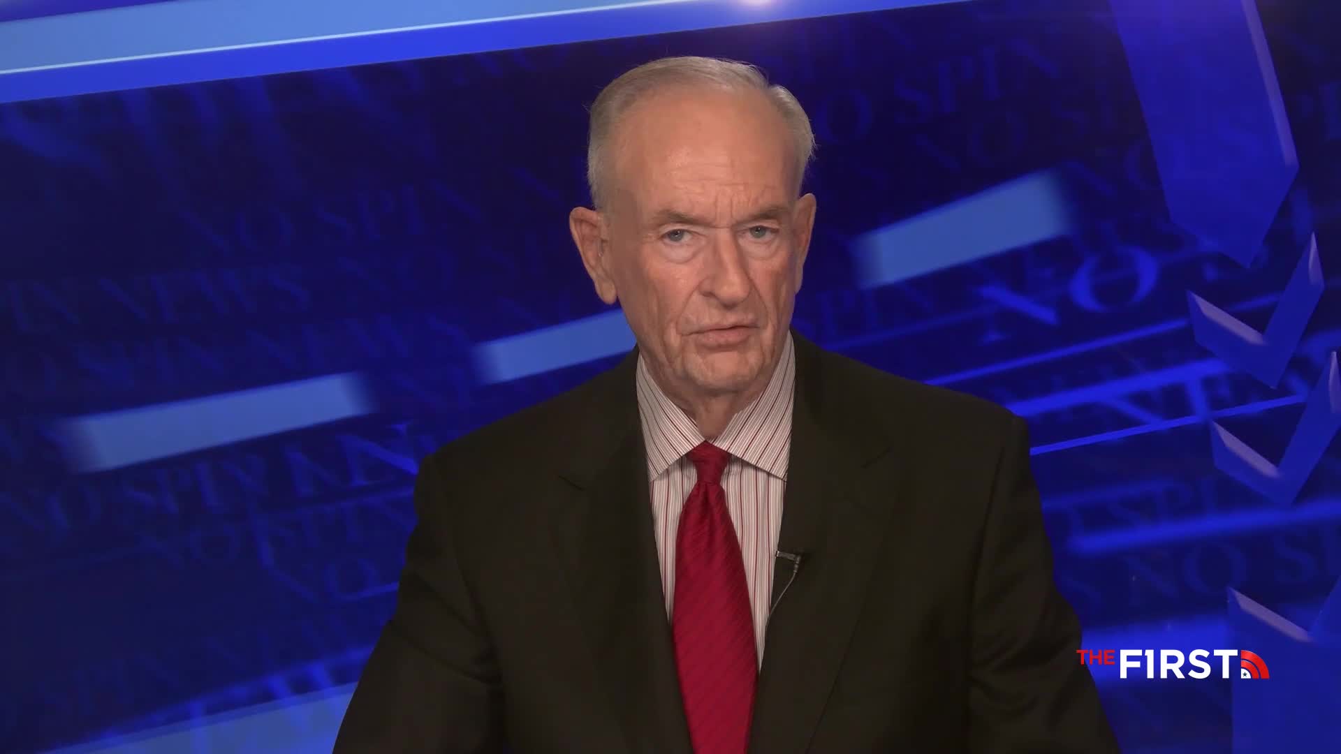 A man in a suit and tie is speaking on a television set, the logo for The First TV visible in the corner. His gaze is directly at the camera, in front of a blue backdrop.
