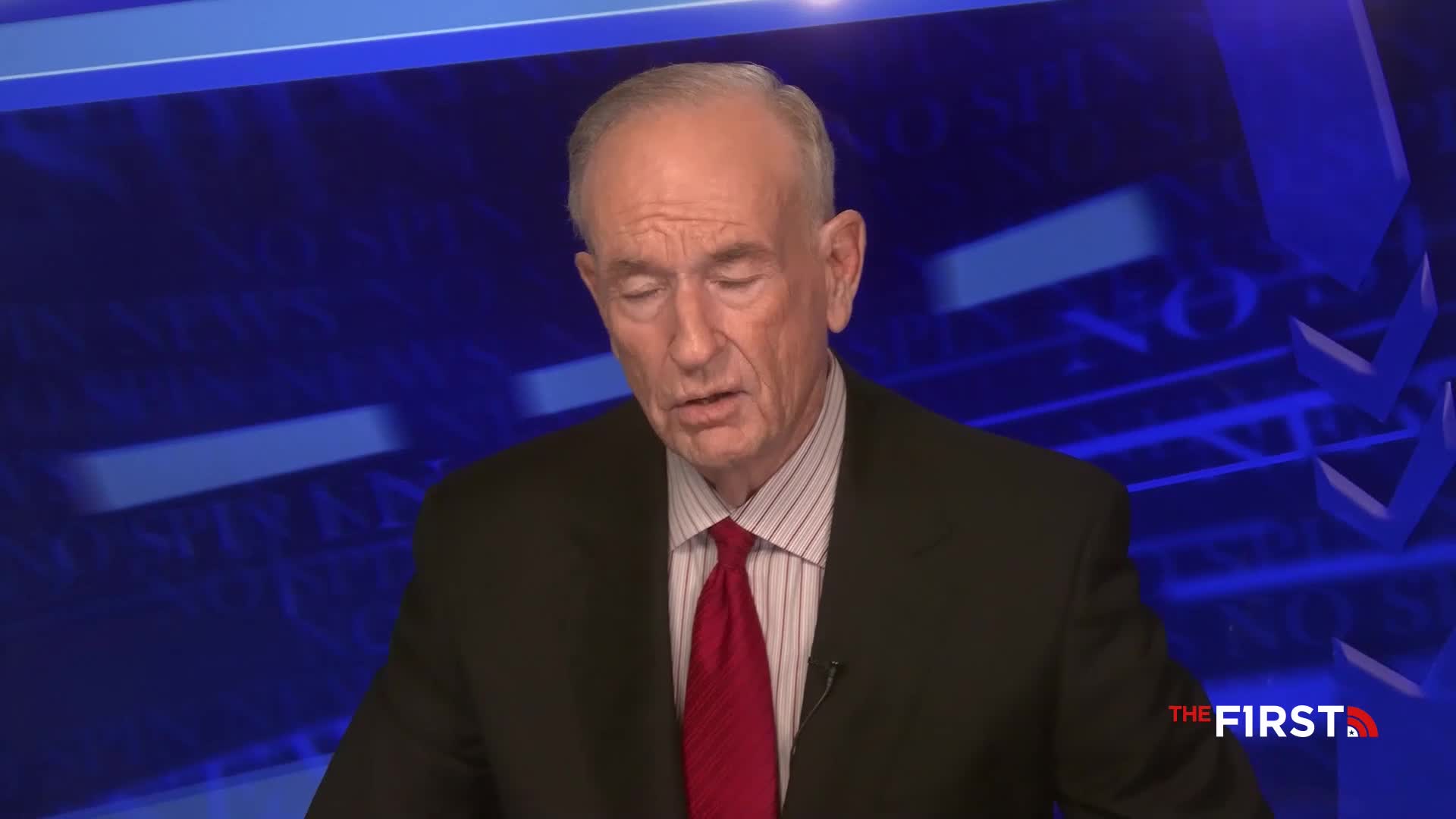 A man in a suit and tie speaks, his gaze directed downward. The logo for The First TV is visible in the corner of the screen.
