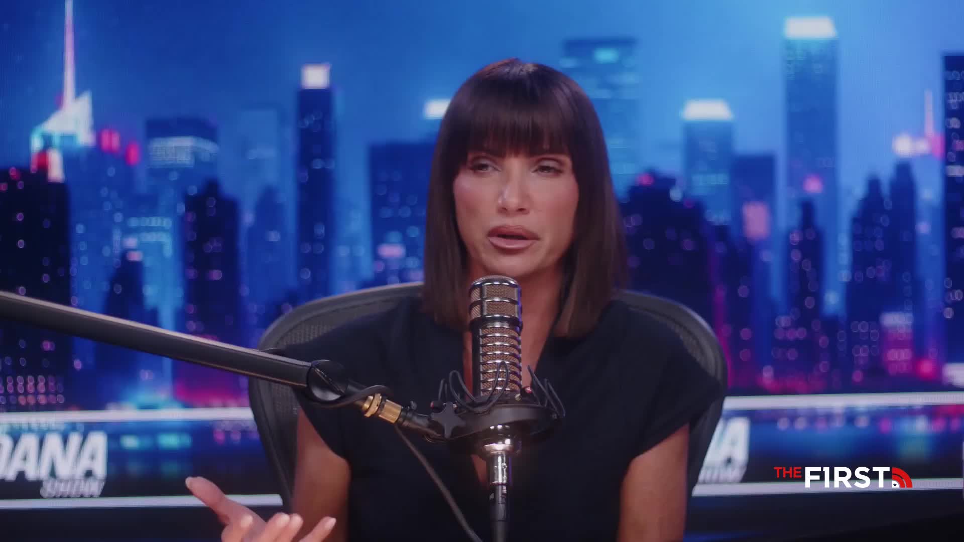 A woman with dark hair sits in front of a microphone, talking on a set for The First TV. Her hands are gesturing as she speaks, in front of a backdrop of a city skyline.
