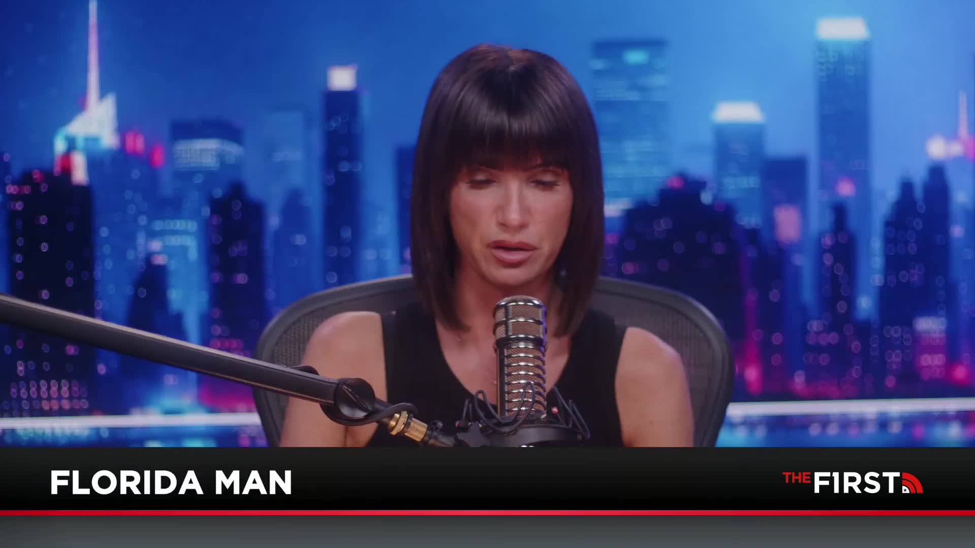 A woman with dark hair sits in a chair, speaking into a microphone. Behind her, a cityscape backdrop glows with the neon lights of a United States city, as seen on The First TV.
