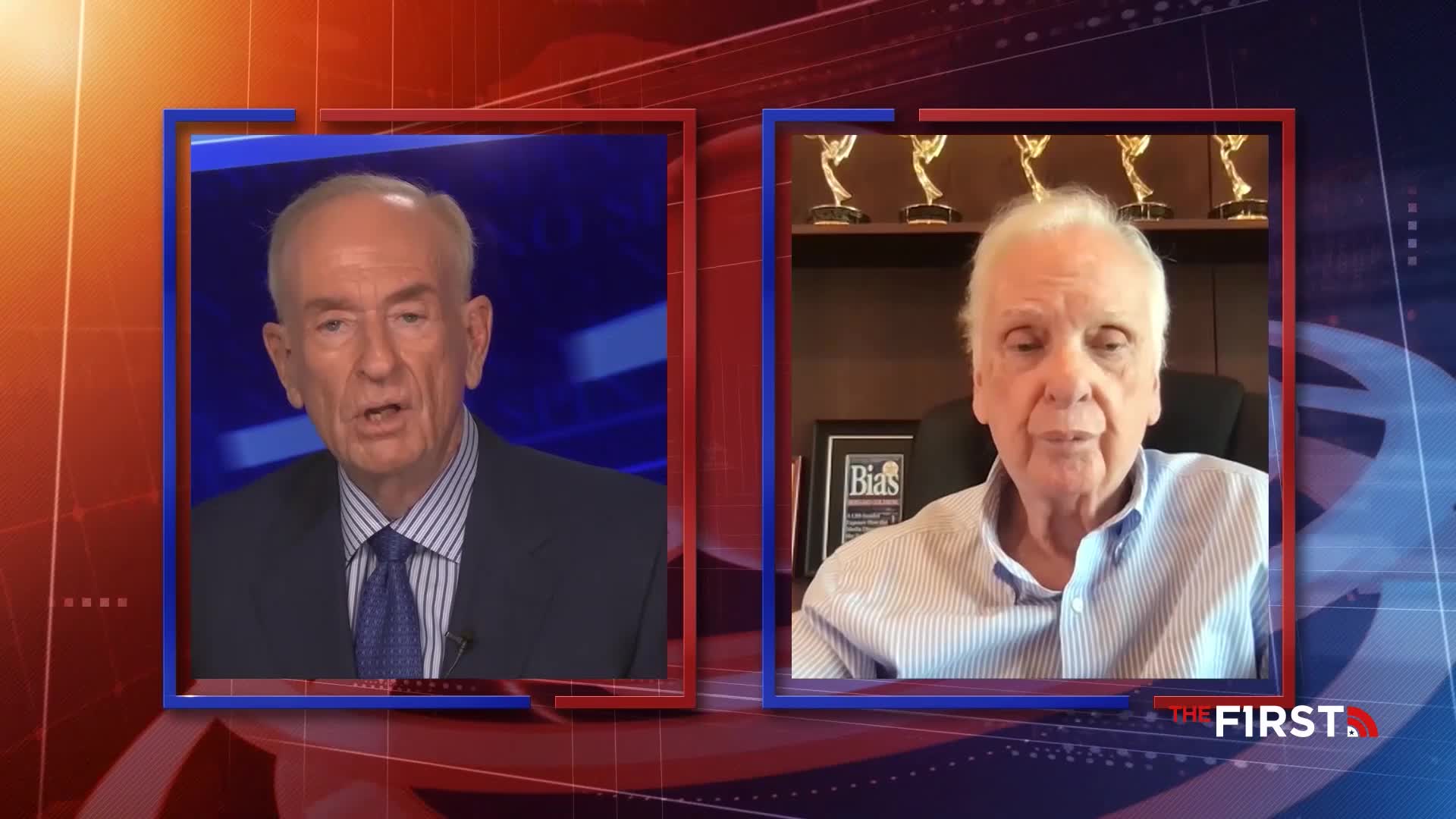 Two men are framed in separate boxes on a TV screen. One, in a suit and tie, speaks from a studio, while the other, in a button-down, appears to be at home, with awards visible behind him.
