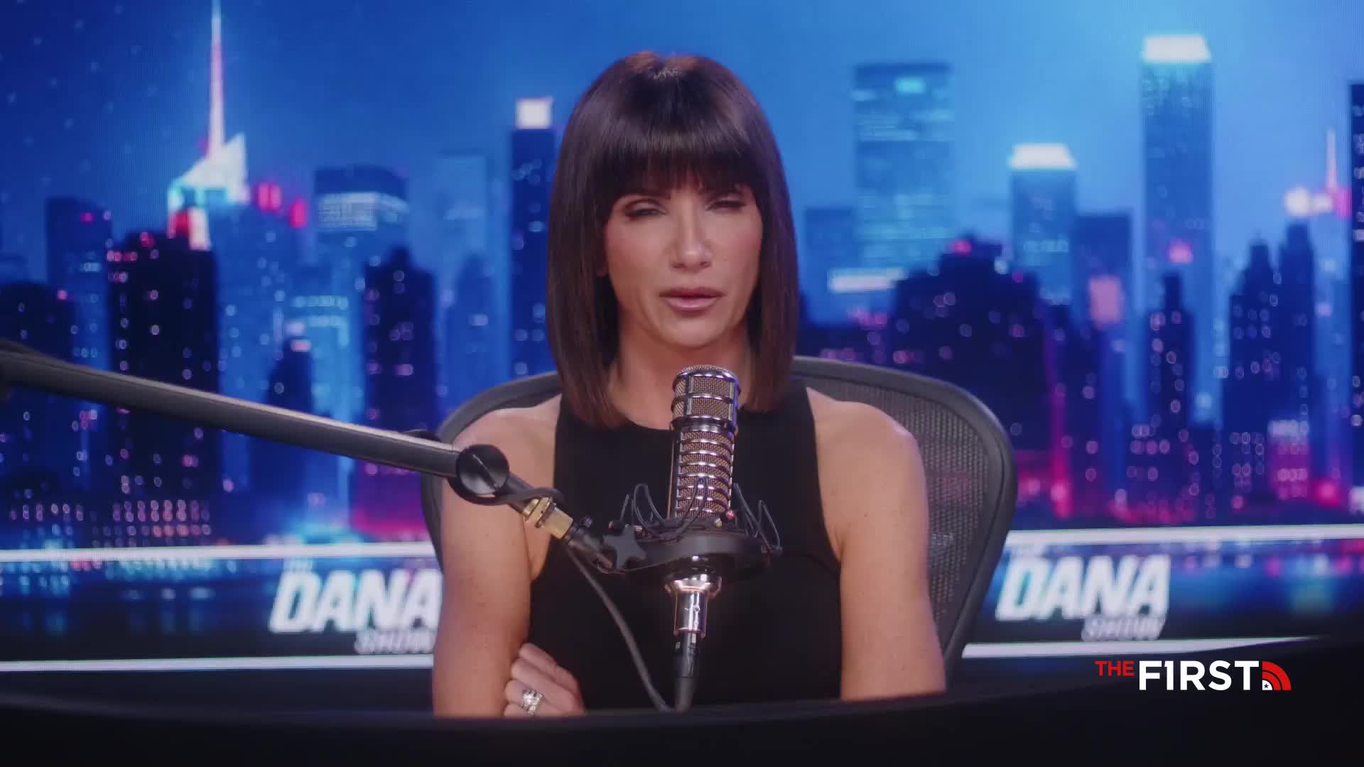 The host of "The Dana Show" on The First TV in the United States sits at a desk, speaking into a microphone. Behind her, a stylized city skyline glows under the studio lights.

