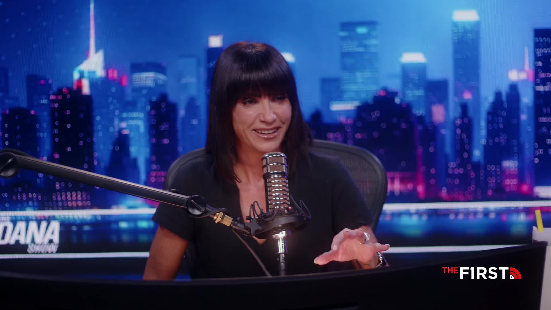 A woman with dark hair gestures with her hands while speaking into a microphone. The backdrop shows a cityscape lit up in blue and pink, and the logo "THE FIRST" is visible on the desk.
