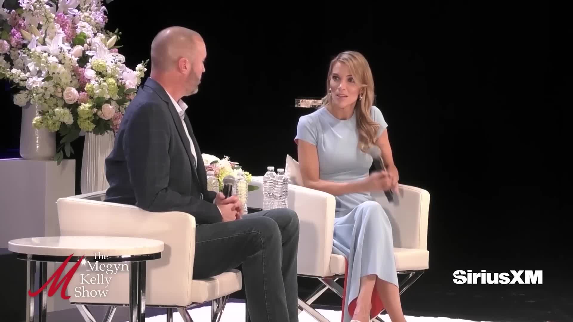 On a stage, Megyn Kelly gestures while speaking, holding a microphone. Beside her, a man in a dark suit listens intently, seated in a white chair.
