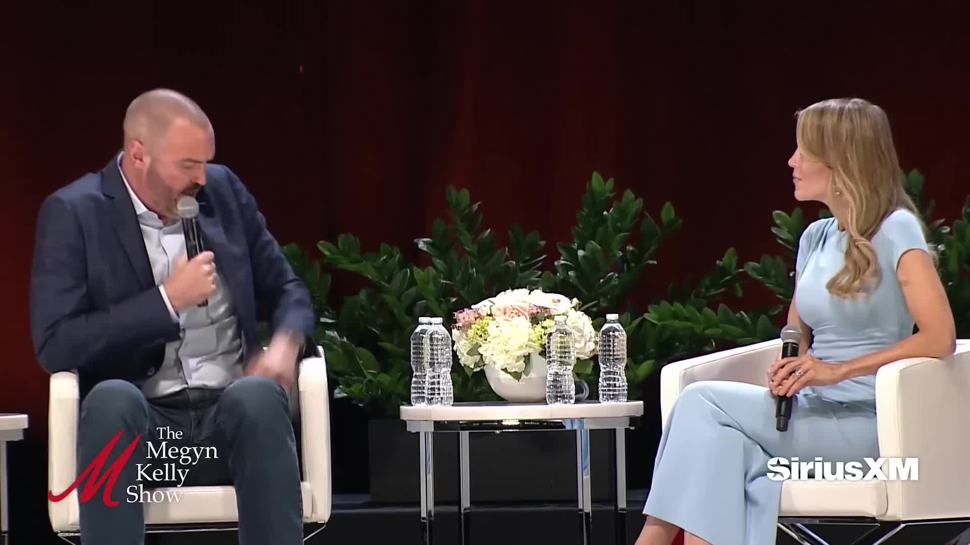 A man in a navy blazer gestures with his left hand while speaking into a microphone, sitting across from a woman in a light blue outfit. They're on a stage, with a table between them holding water bottles and a floral arrangement.
