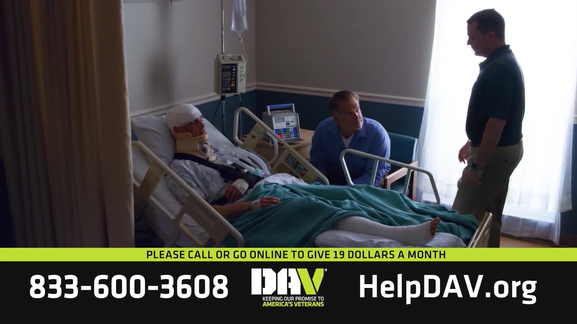 A man lies in a hospital bed, his head and neck braced, his leg in a cast. Two men stand beside him, one leaning in to speak.
