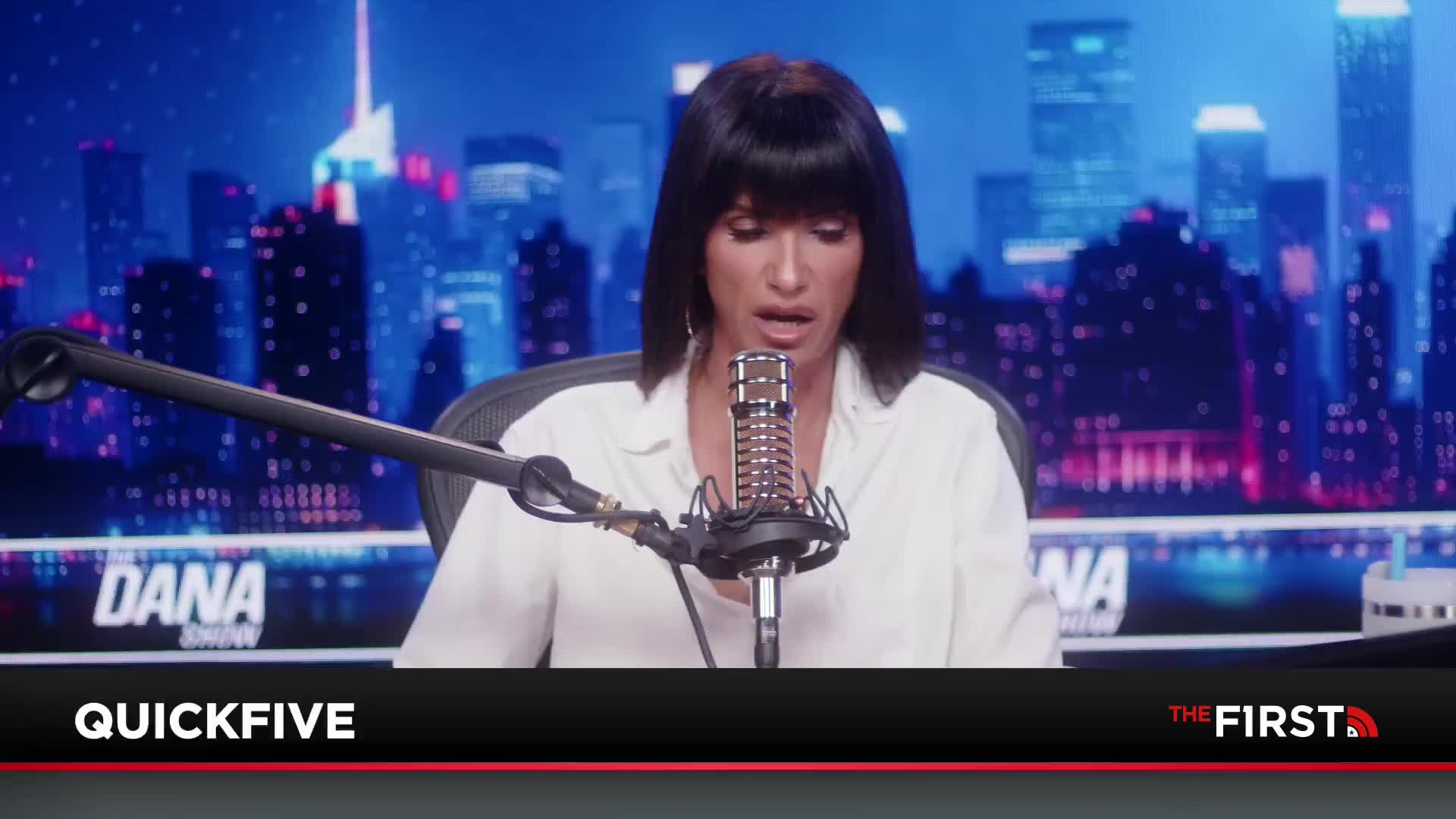 The host of "The Dana Show" on The First TV is speaking into a microphone, the New York City skyline a backdrop behind her. Her dark hair frames her face as she appears to be discussing something with a serious expression.
