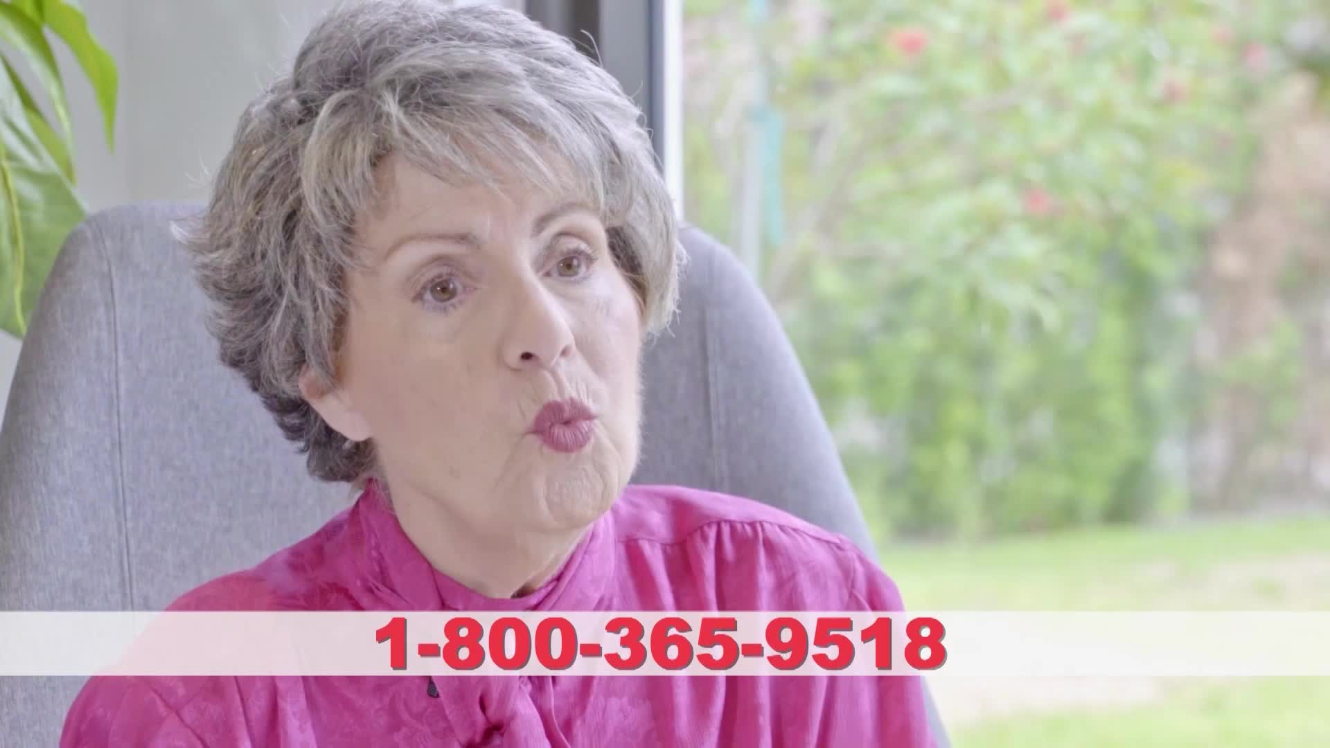 A woman with gray hair, wearing a pink shirt, sits in a chair and speaks. A phone number appears at the bottom of the screen.
