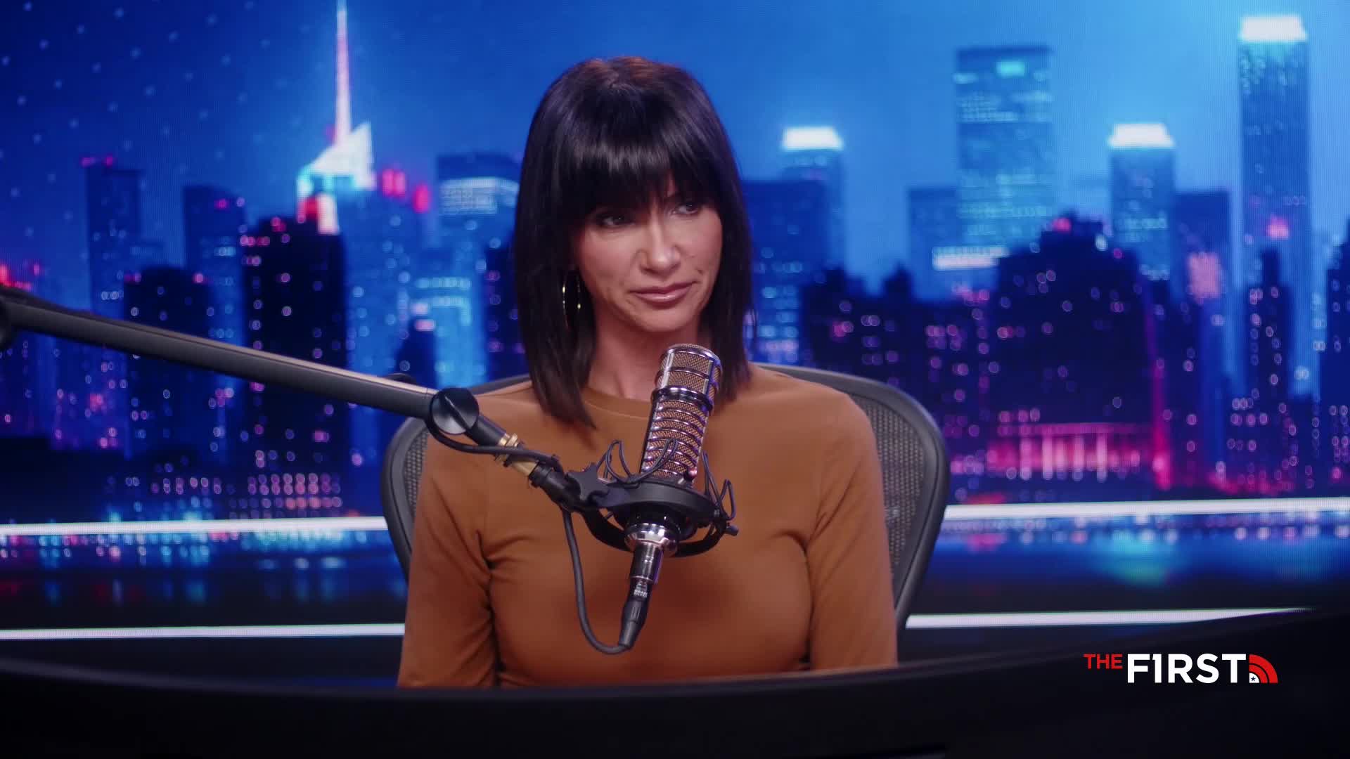 A woman with dark hair sits in front of a microphone, speaking into it. Behind her, a cityscape backdrop glows with blue and white lights, and the logo for "The First" is visible.
